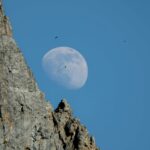 Stunning view of the moon rising above jagged mountain formations under a clear blue sky.