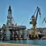 An offshore oil rig surrounded by cranes in a sunny industrial harbor setting.